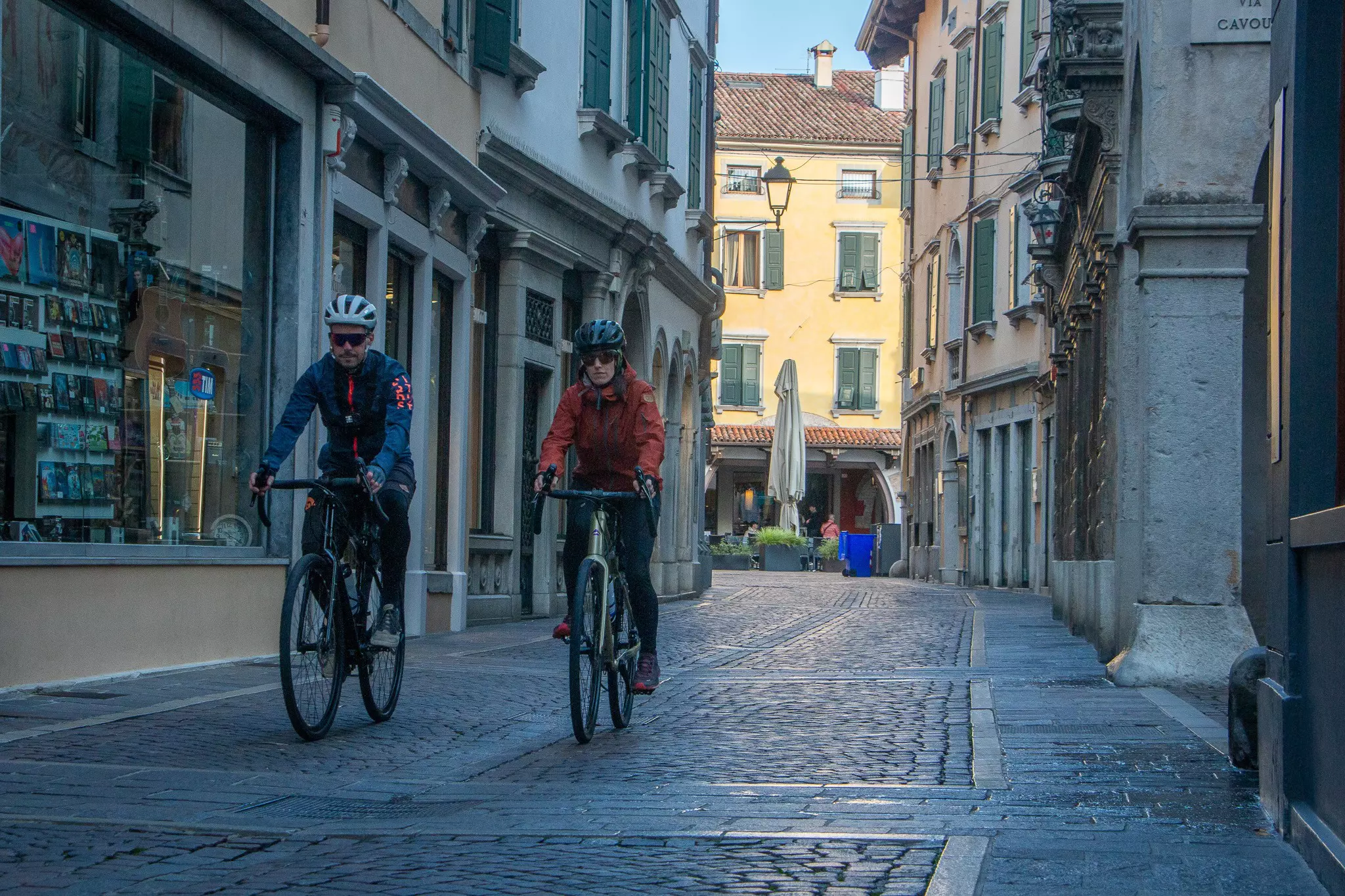 Two cyclists pedal through a quaint town with cobbled streets.