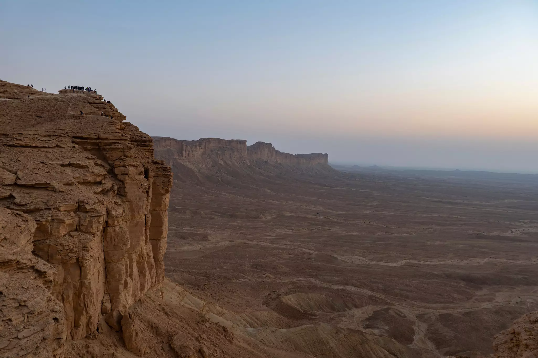 Small figures are clustered at the edge of a cliff at sunset.