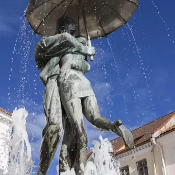 Bronze statue of a couple embracing under an umbrella, water spraying around them against a deep blue sky.