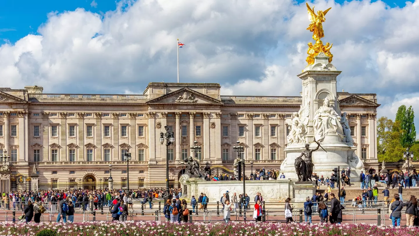 Tourists wander near a large stately home with an ornate gold-topped statue in front of it. Nearby flowerbeds are packed with pink blooms.