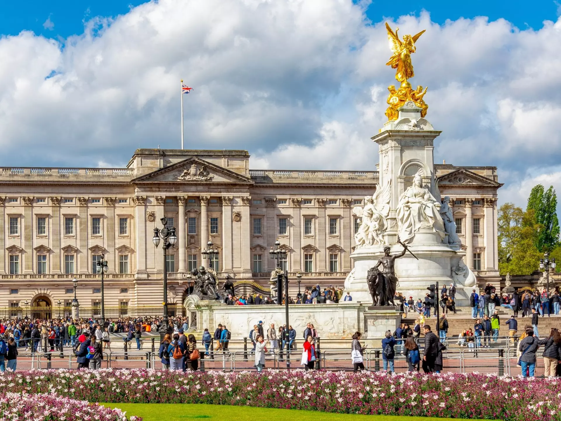 Tourists wander near a large stately home with an ornate gold-topped statue in front of it. Nearby flowerbeds are packed with pink blooms.