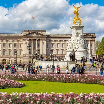 Tourists wander near a large stately home with an ornate gold-topped statue in front of it. Nearby flowerbeds are packed with pink blooms.
