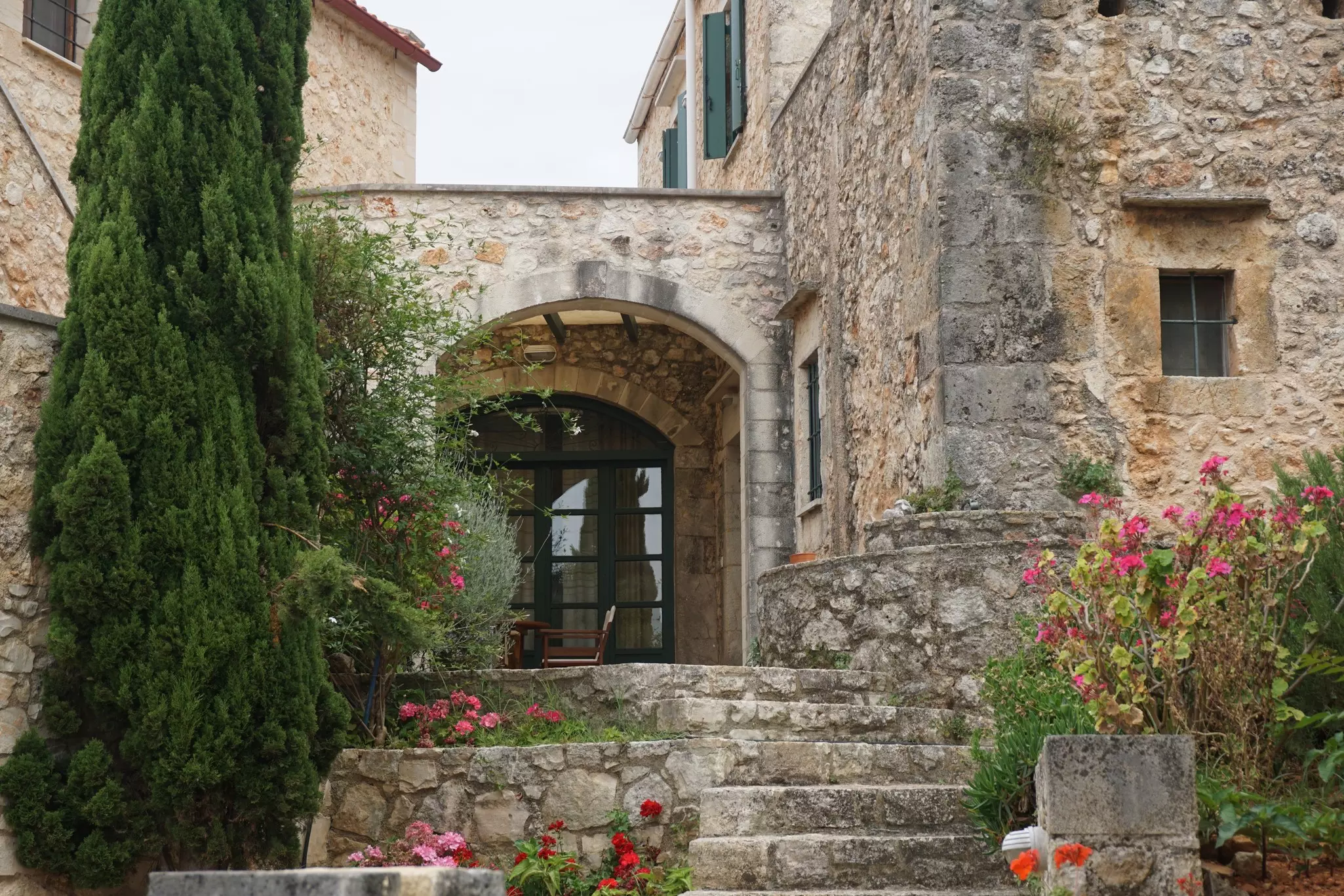 Steps lead to the arched entryway of a stone building. Flowers and a cypress tree are seen beside the steps.