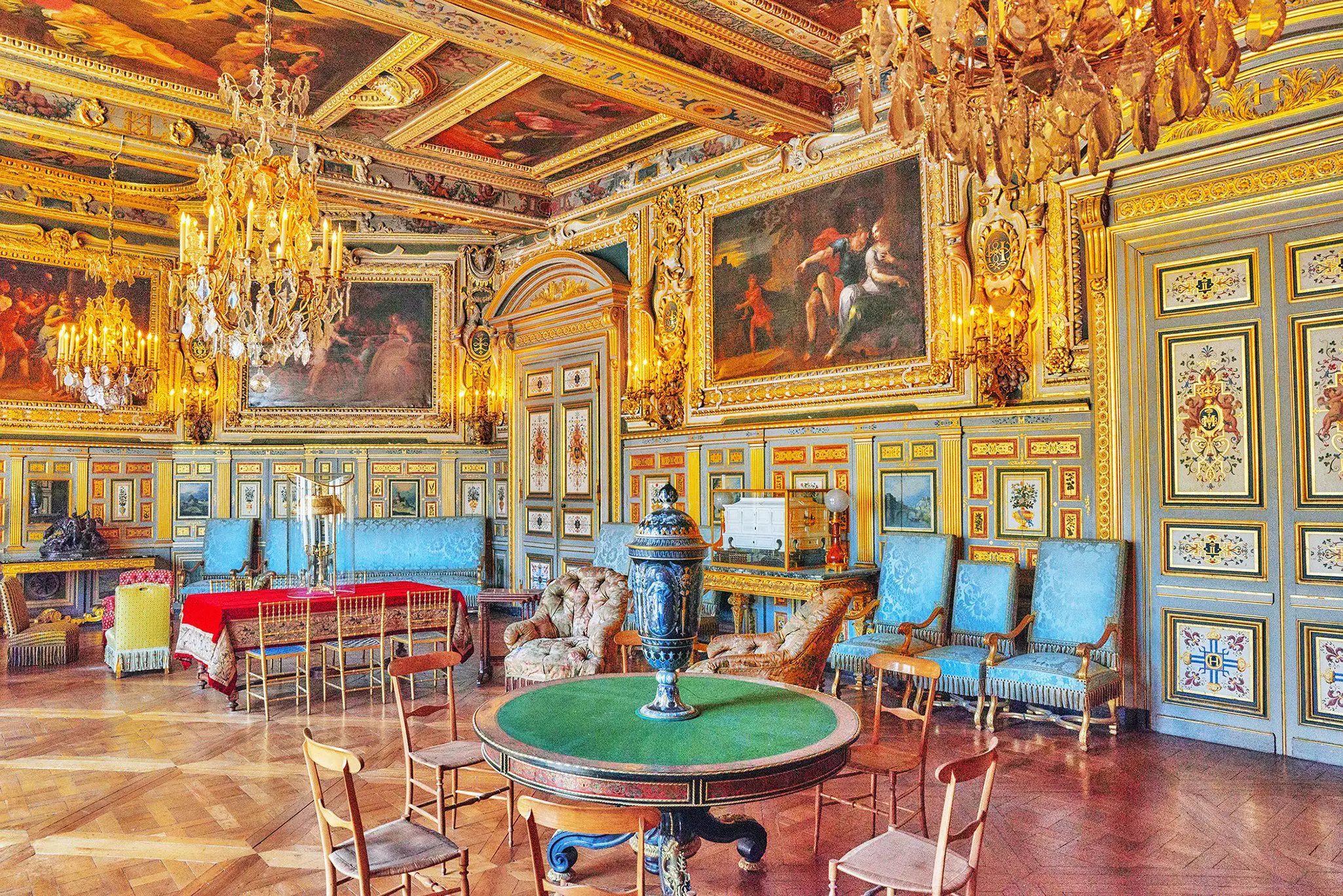 An opulent palace room, with chandeliers, gold paint, large oil paintings and a lot of chairs.