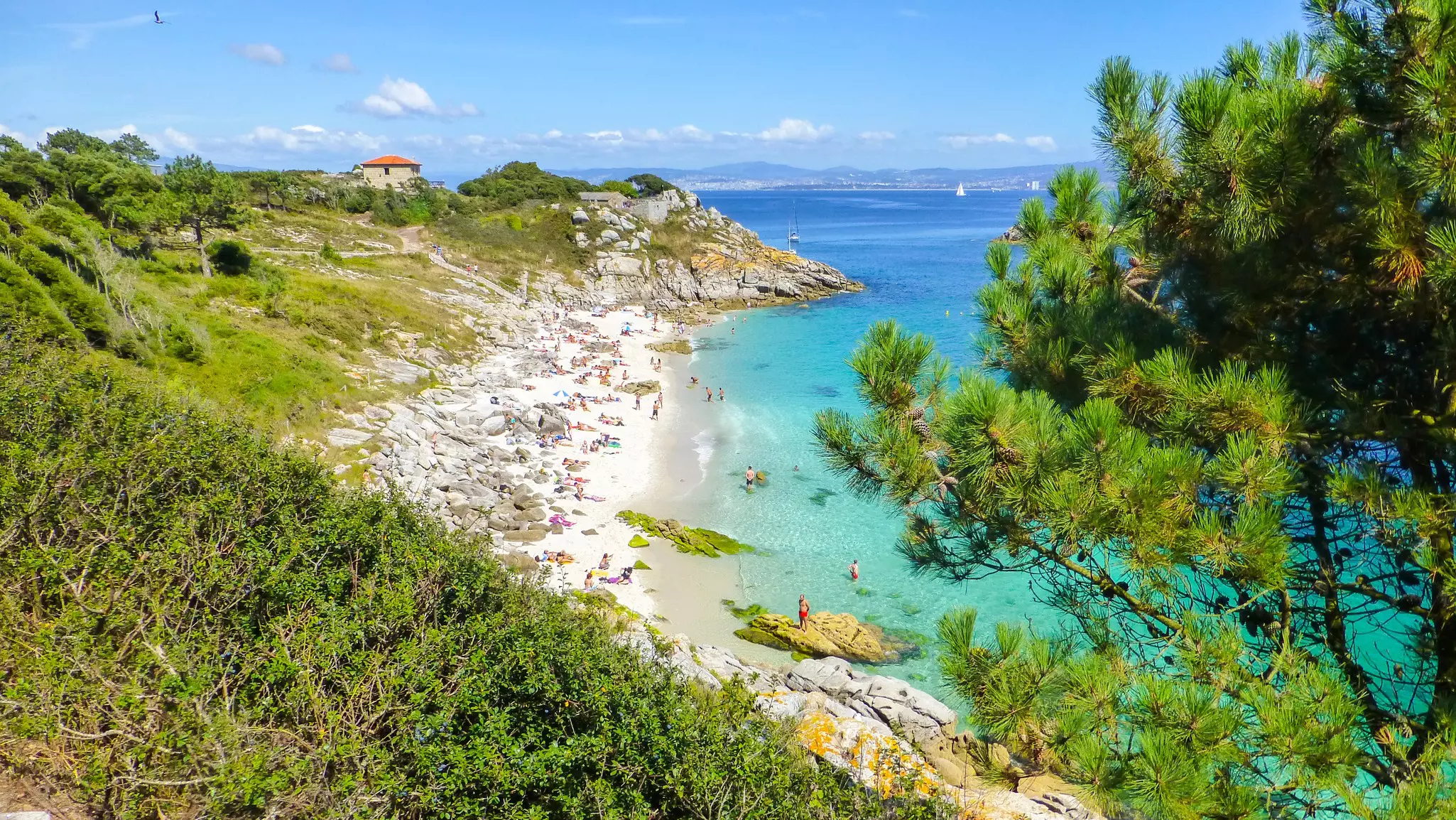 People at a sandy cove against a green hillside facing turquoise water.