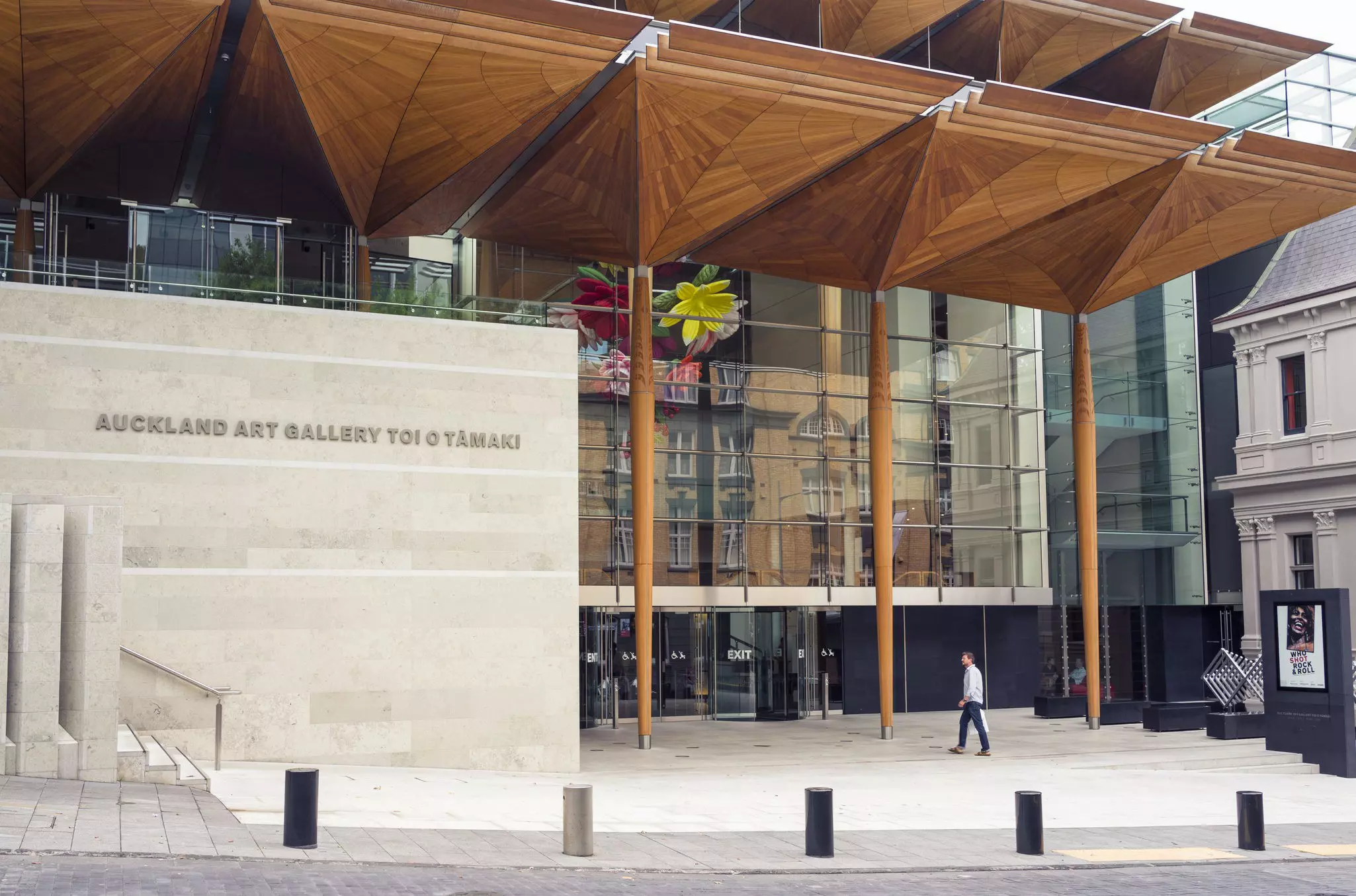 A man walks on concrete walkway toward the exterior of a building of stone and glass.
