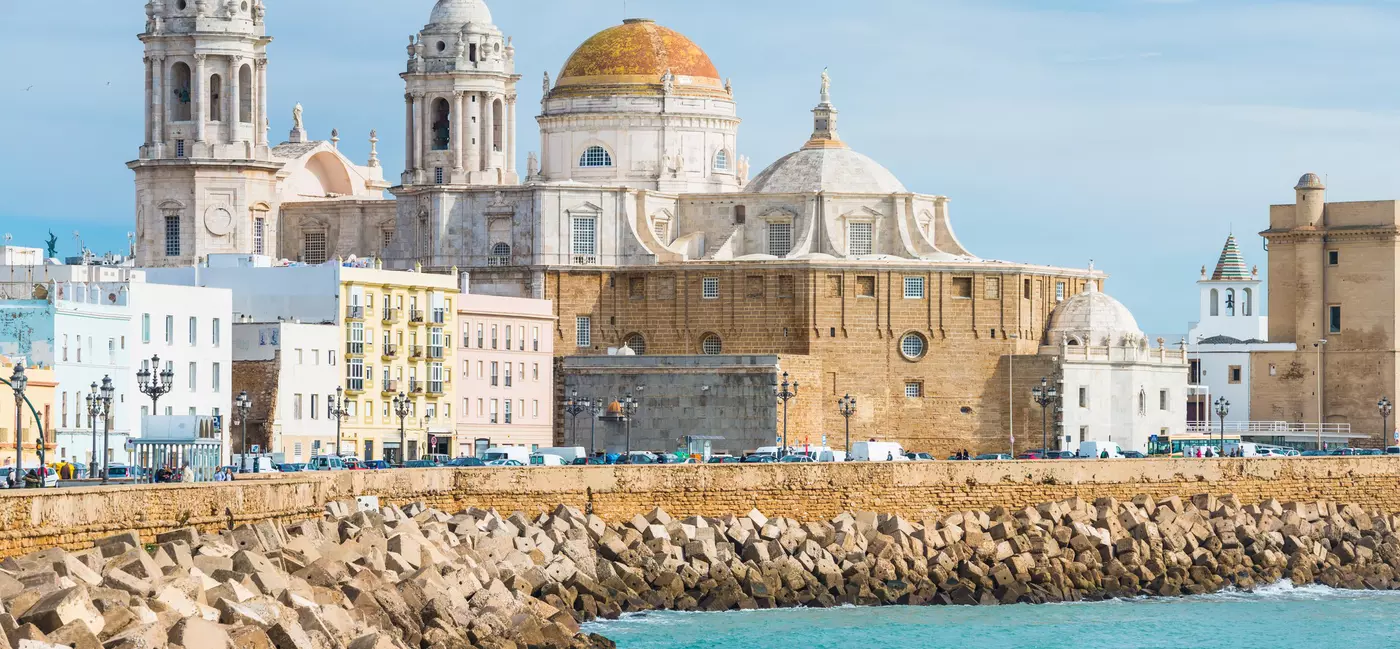 A cathedral with a bronze dome roof and white towers overlooks a rocky embankment by the sea.