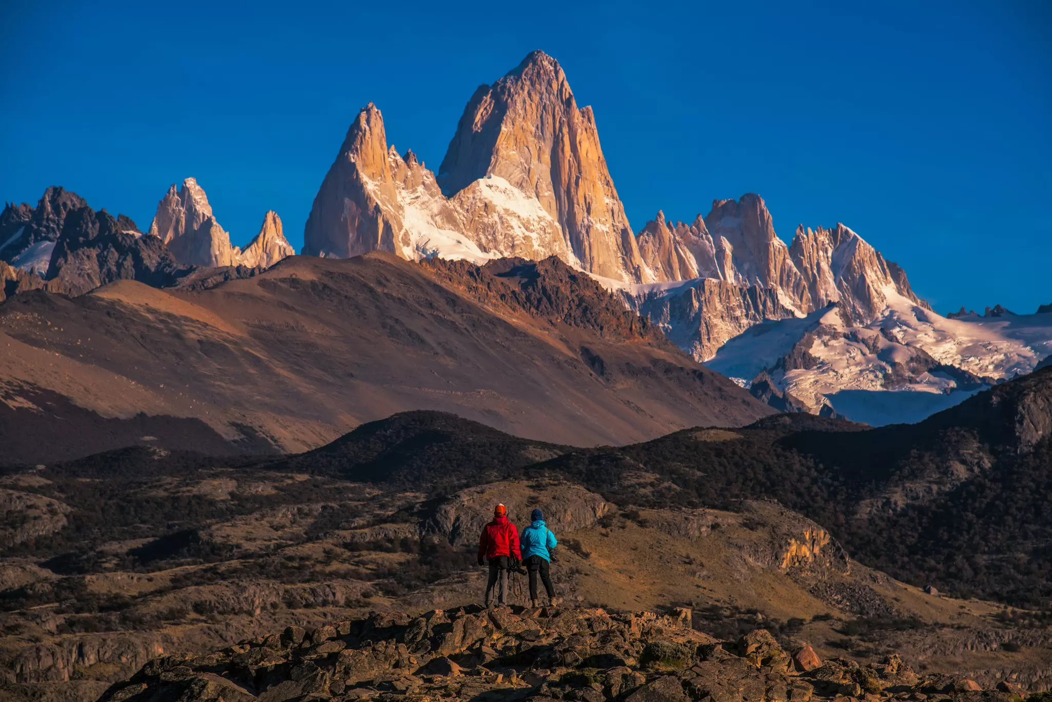 A couple stand with their backs to the camera and look over a parched landscape and up to the highest summits of Los Glaciers National Park.