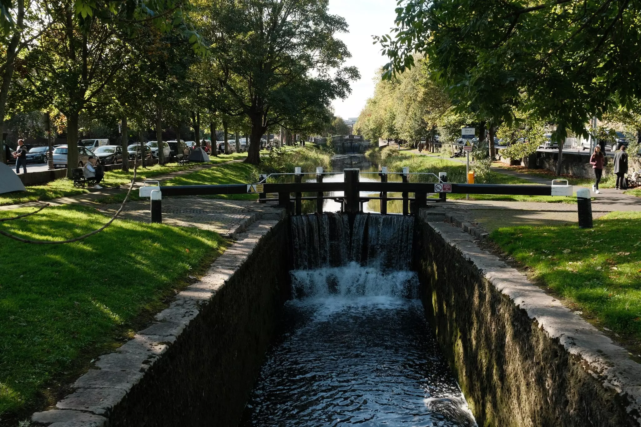 A small lock in a narrow canal lined by grassy banks in a city.