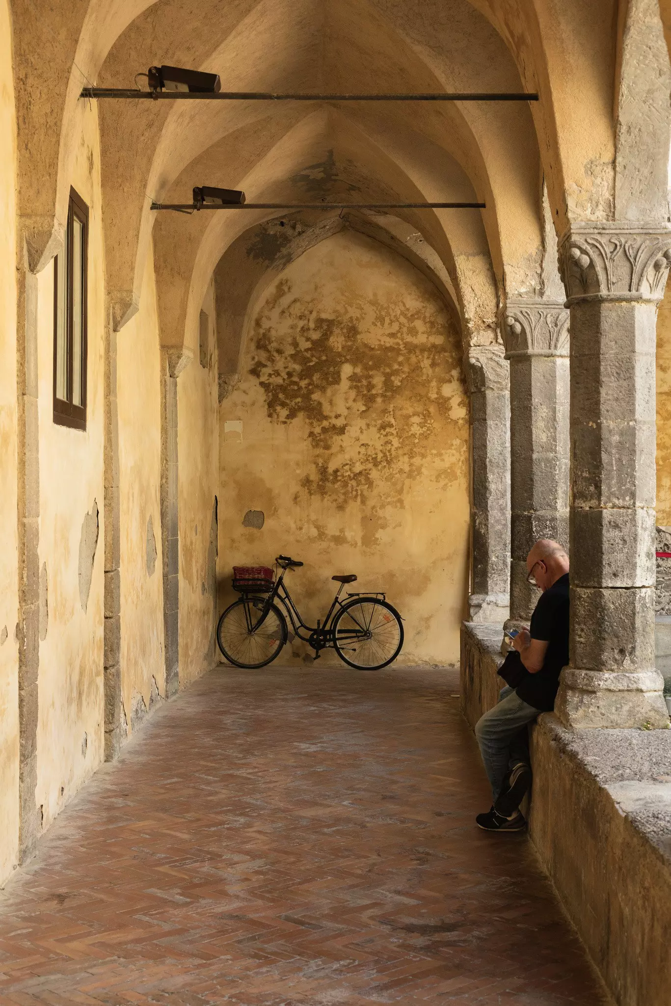 A man leans along a column in a stone courtyard with a parked bicycle in the far distance.