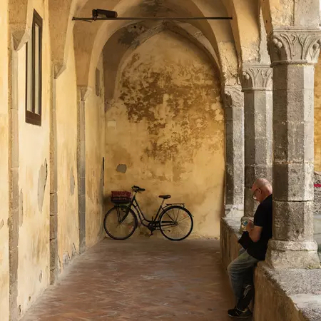 A man leans along a column in a stone courtyard with a parked bicycle in the far distance.
