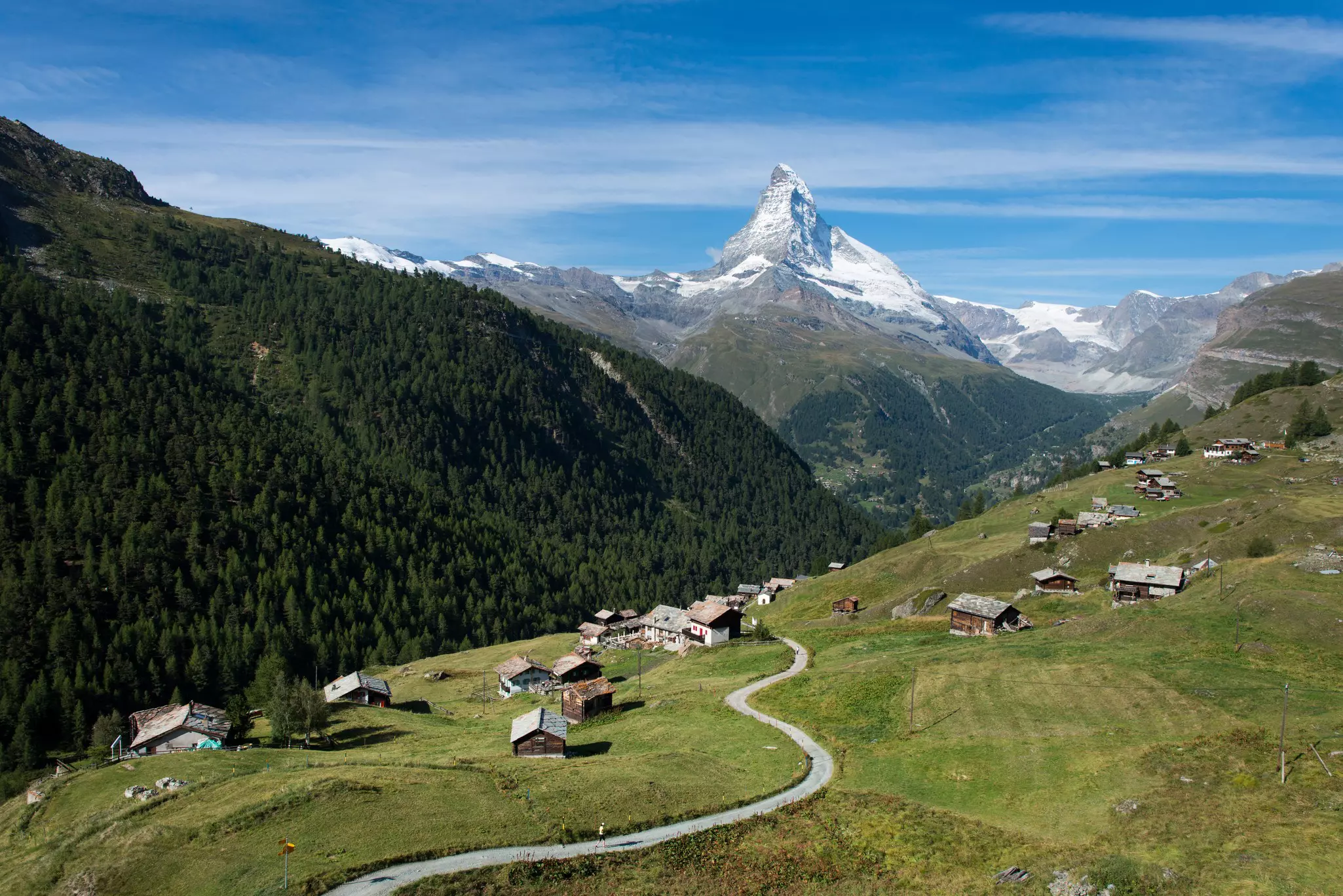 An aerial view of a road on a hillside leading through a mountain village. A huge peak is visible in the distance.