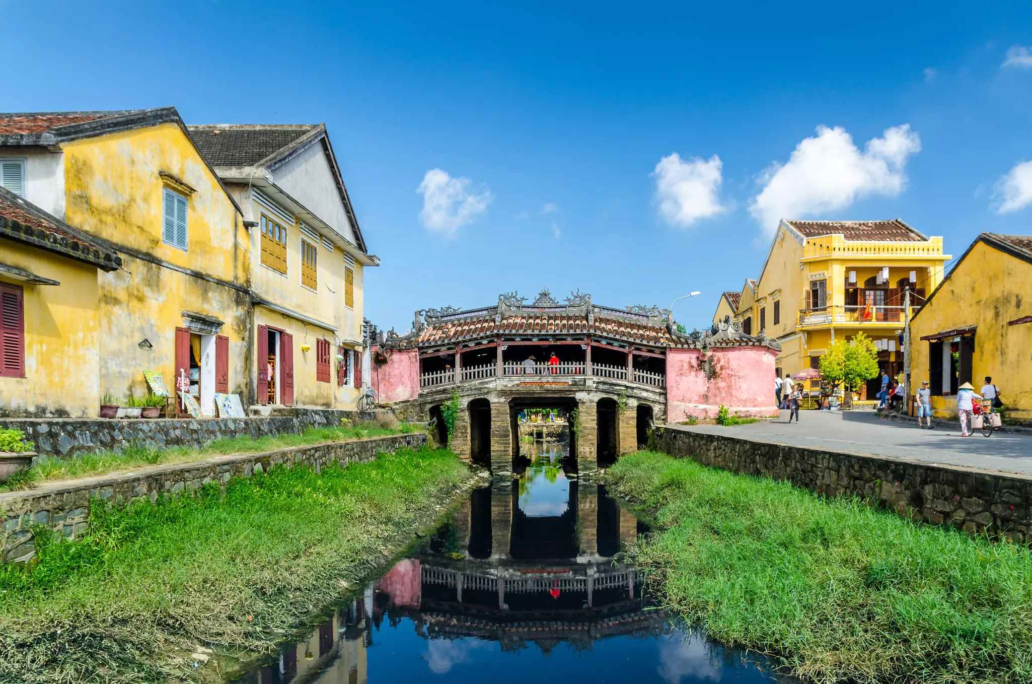 The Japanese Bridge in Hoi An, with water and grass in front of it and yellow buildings nearby