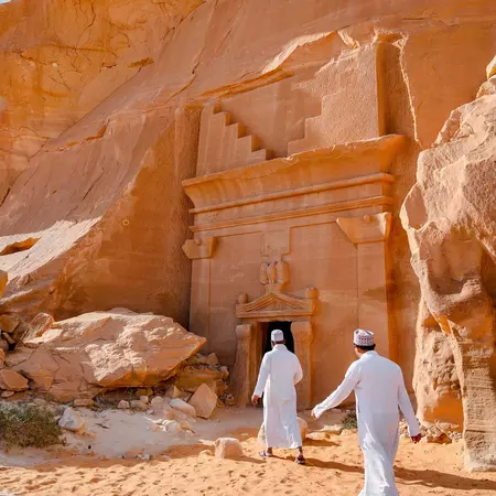Two visitors wearing white walk toward the entrance to a tomb carved into a sandstone rock.