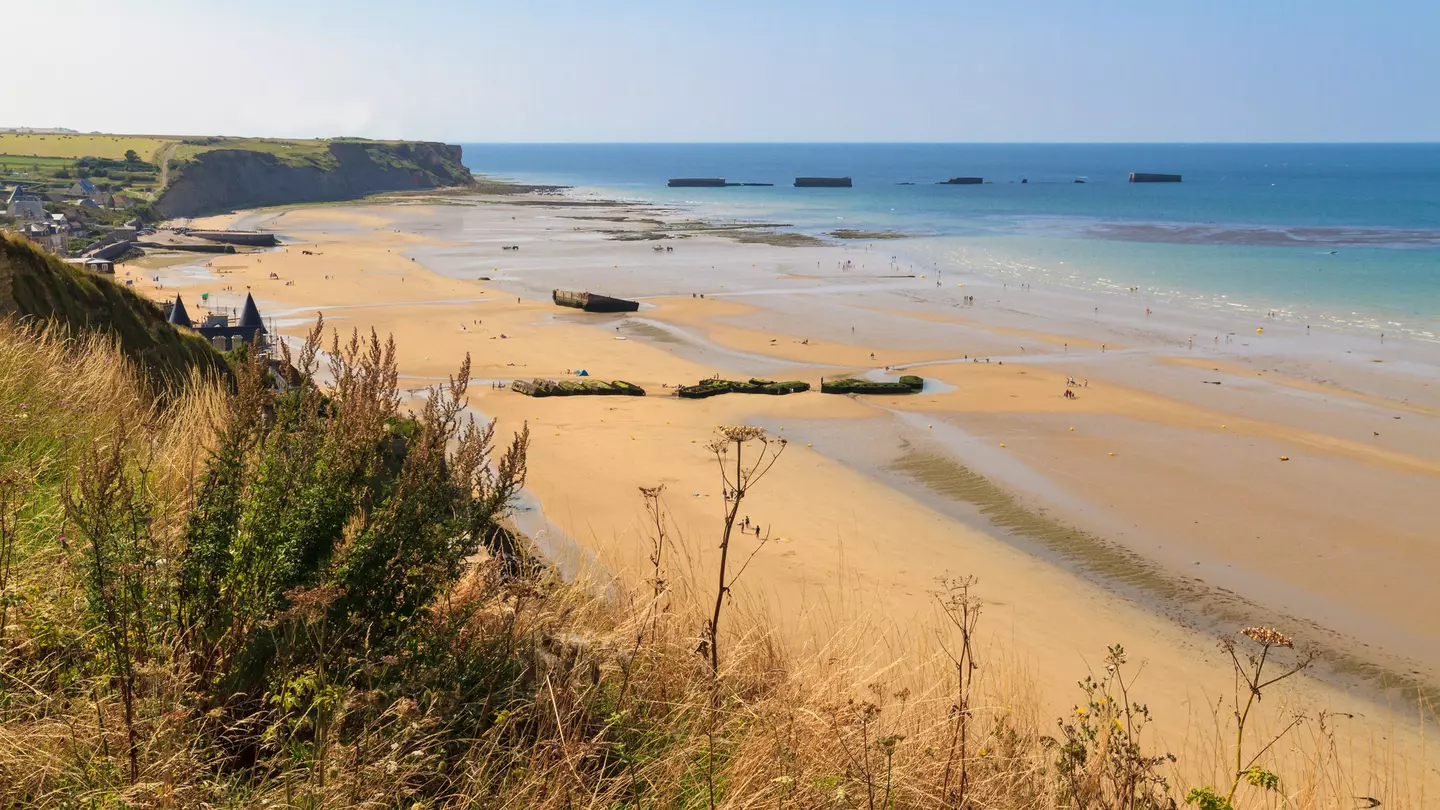 Looking down from a grassy clifftop over a huge golden D-Day landing beach, with the remains of a Mulberry Harbour (artificial port) at Arromanches-les-Bains visible in the distance