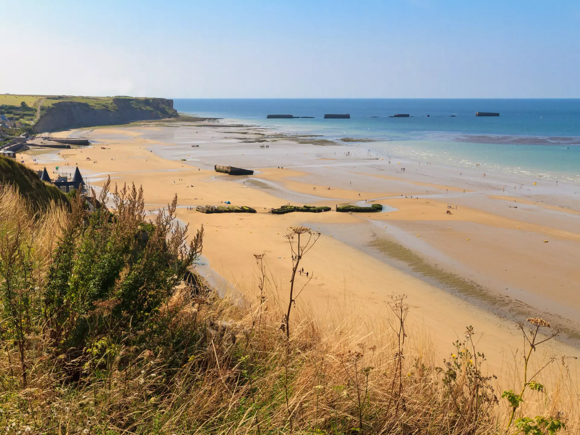 Looking down from a grassy clifftop over a huge golden D-Day landing beach, with the remains of a Mulberry Harbour (artificial port) at Arromanches-les-Bains visible in the distance