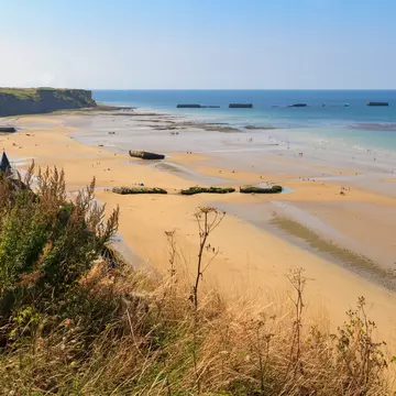 Looking down from a grassy clifftop over a huge golden D-Day landing beach, with the remains of a Mulberry Harbour (artificial port) at Arromanches-les-Bains visible in the distance