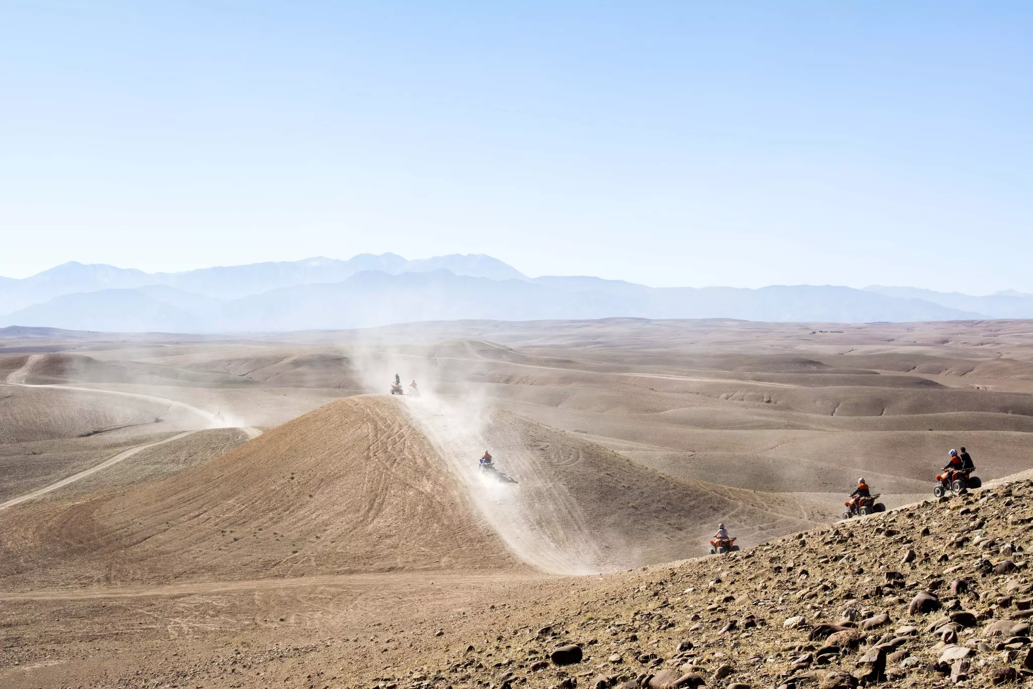 People ride on quad bikes along a dusty track over sand dunes in a vast desert.