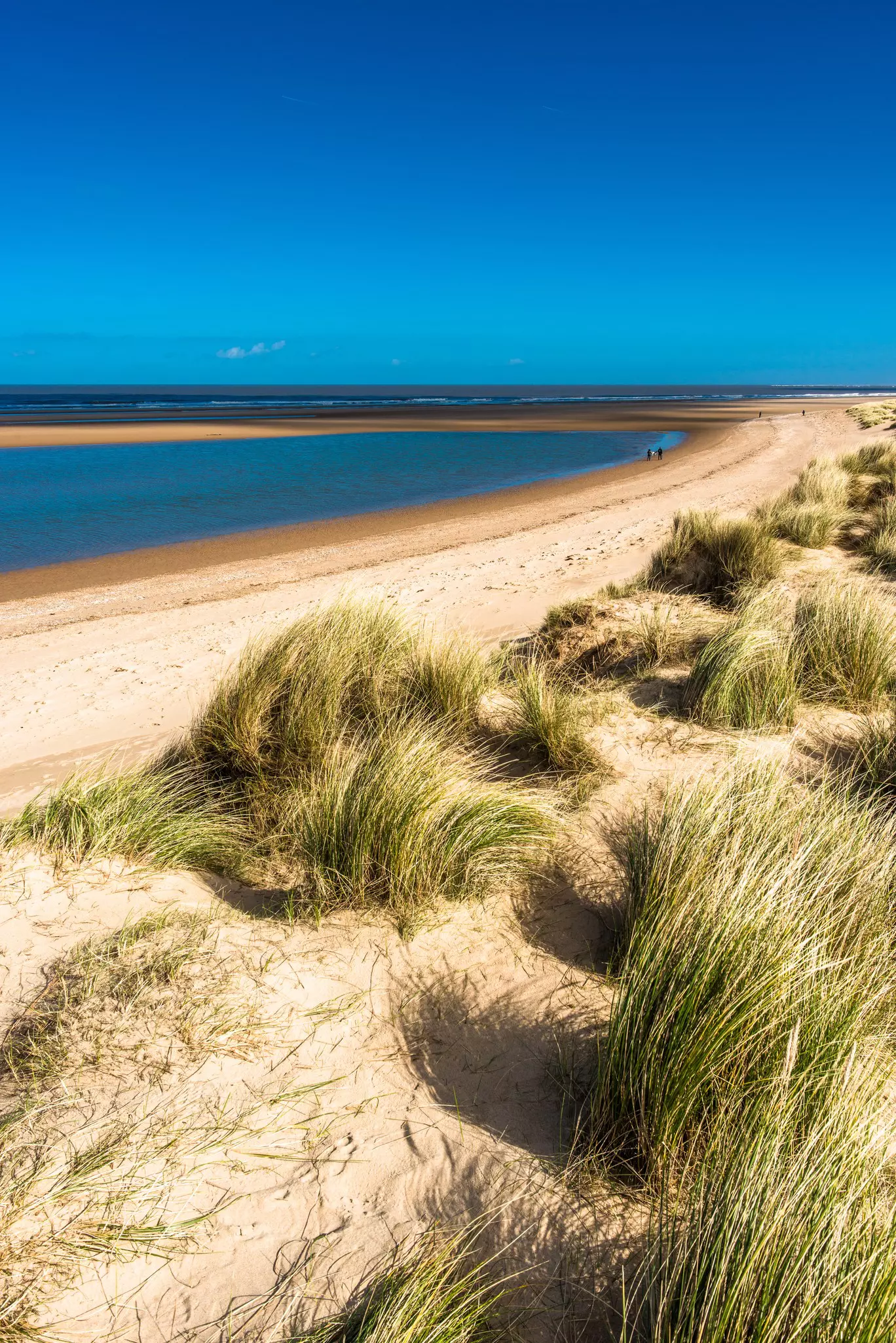 Grass-topped sand dunes above a long stretch of golden sand where two people are walking in the distance