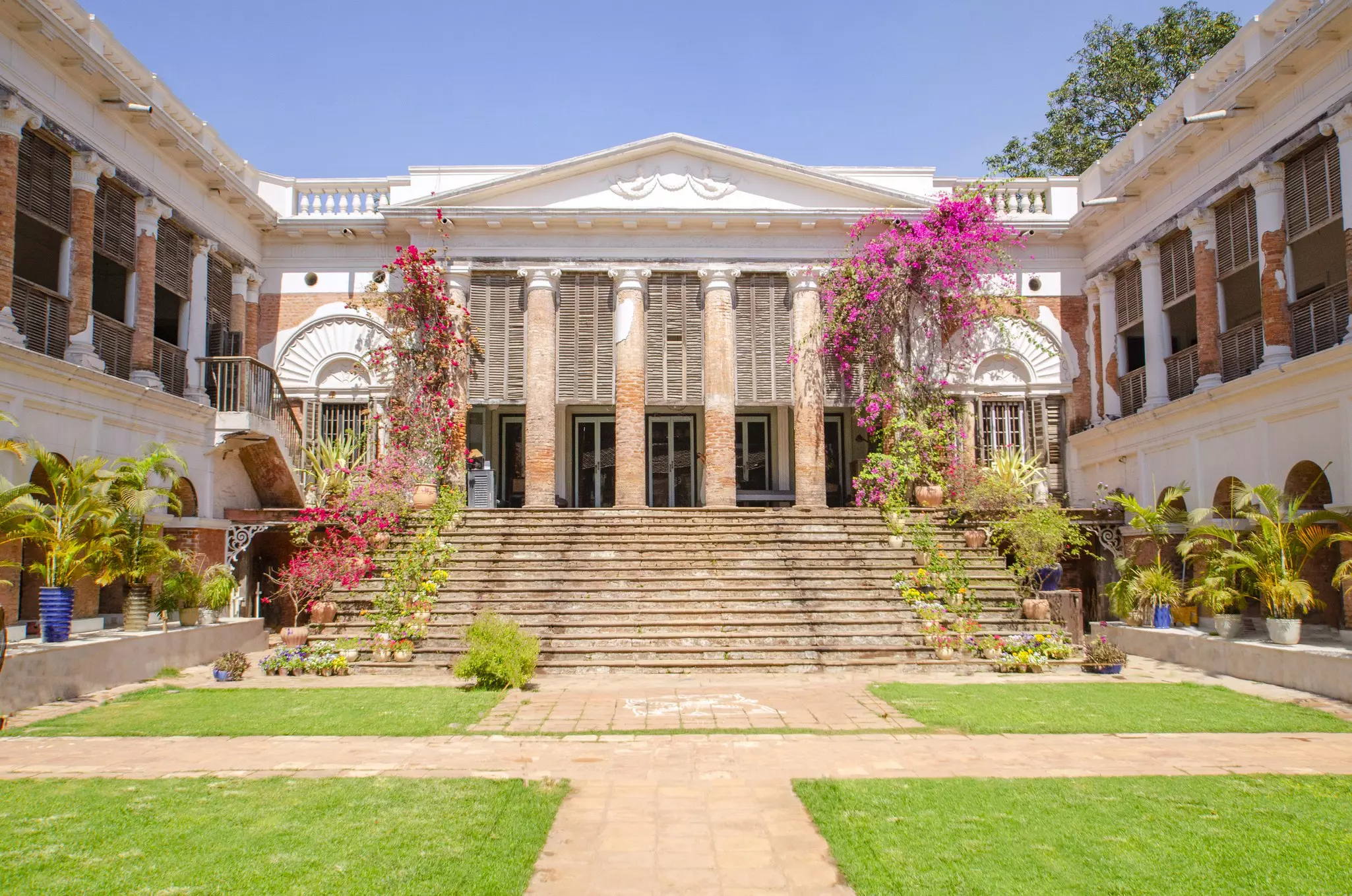 A well-tended courtyard garden gives way to a flight of stone steps leading up to a columned facade of a grand building.