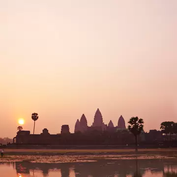Silhouette of Angkor Wat's towers and palm trees at dawn, reflected in still water under a hazy pink sky.
