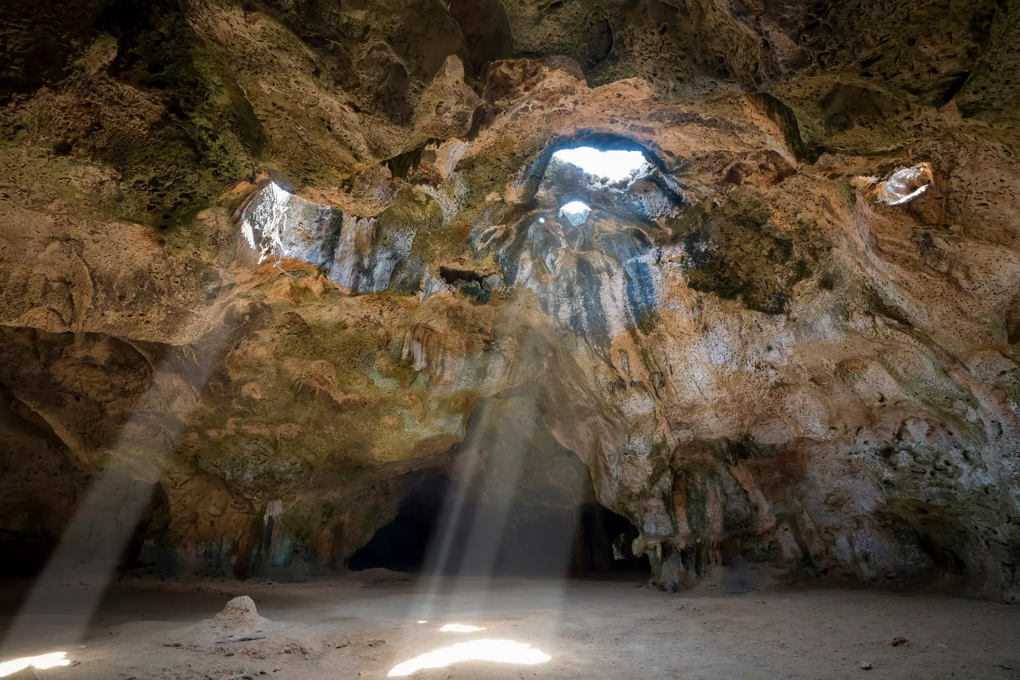 Light streams through openings in a rock cave in Aruba to shine on white sand.