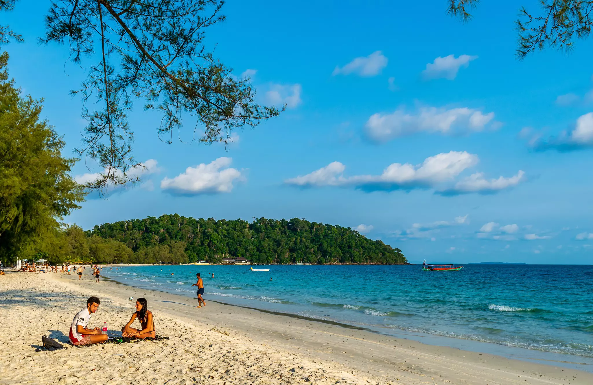 People sit on white sand beach in Cambodia next to blue ocean; in the background, a few people are walking along the shore. There is a boat in the water, and vegetation on the other side of the cove.