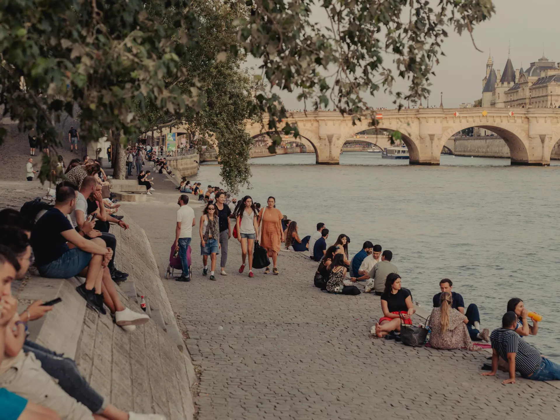People sitting by a river, with an arched bridge in the background.