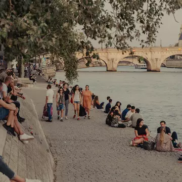 People sitting by a river, with an arched bridge in the background.