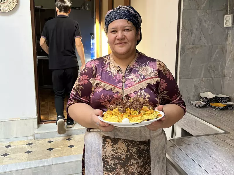 A woman in a head covering stands in kitchen holding a big dish of plov, and Uzbek rice dish.