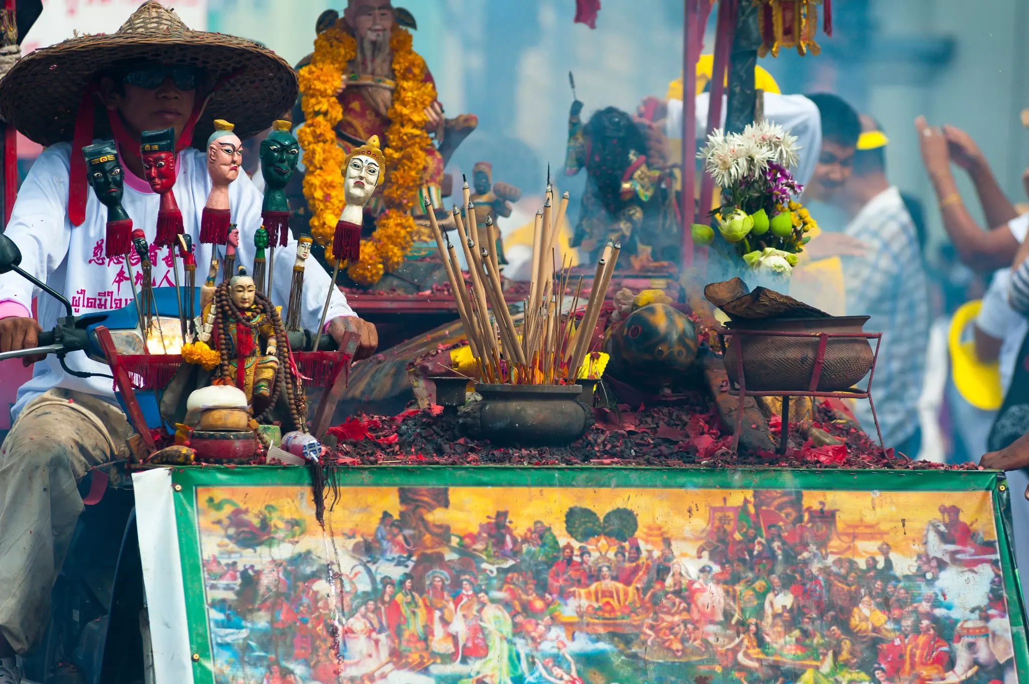A vendor in a straw hat sits behind a table with a colorful painting in front and various wares, such as figures on sticks, with flower petals strewn about. People celebrate in the background.