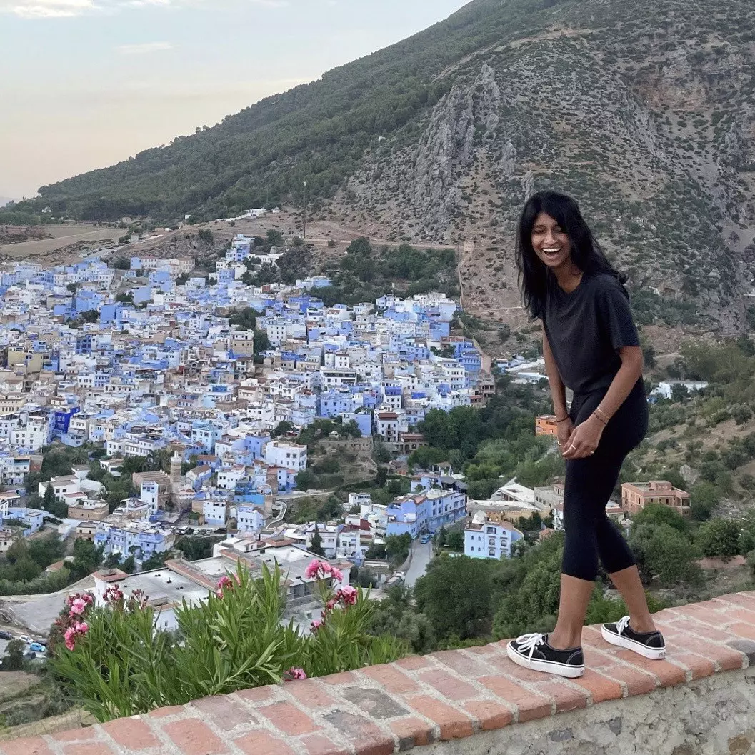 Deepa Lakshmin in Morocco, laughing and standing on a wall with the city of Chefchaouen in the background