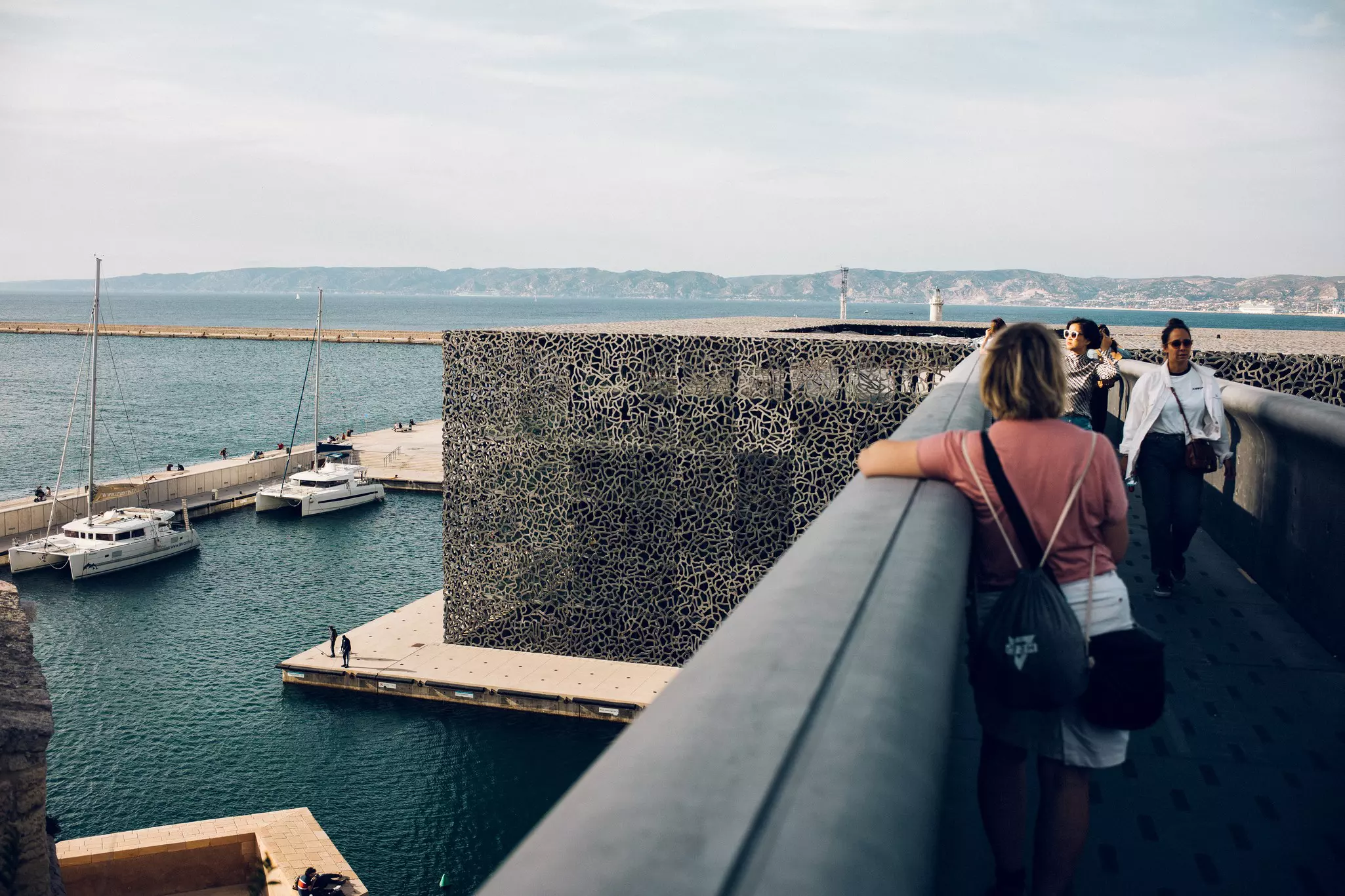 People are seen on a walkway above a large building by the port of a city.