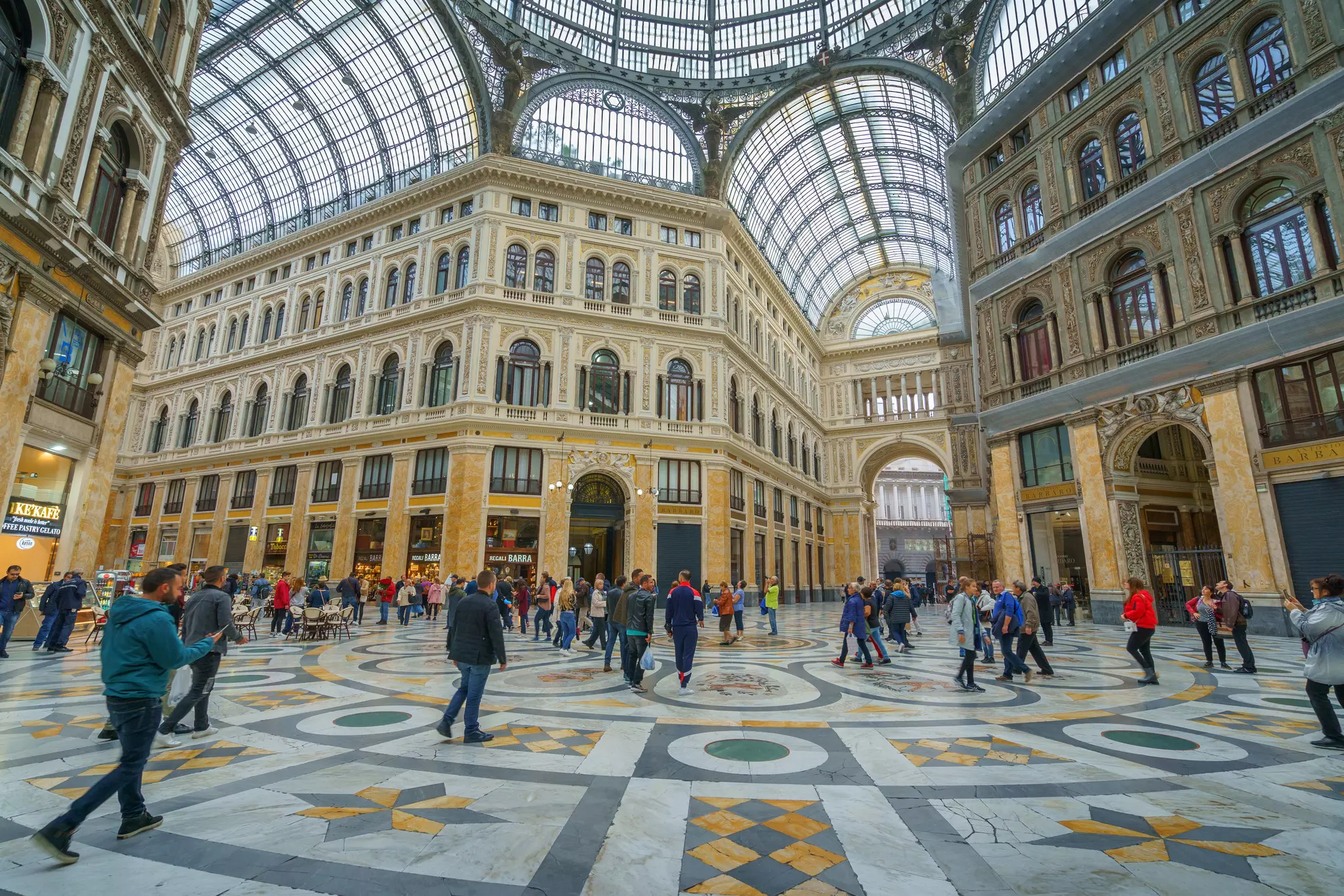 People walk in a shopping arcade with marble tiles and a vaulted glass ceiling.