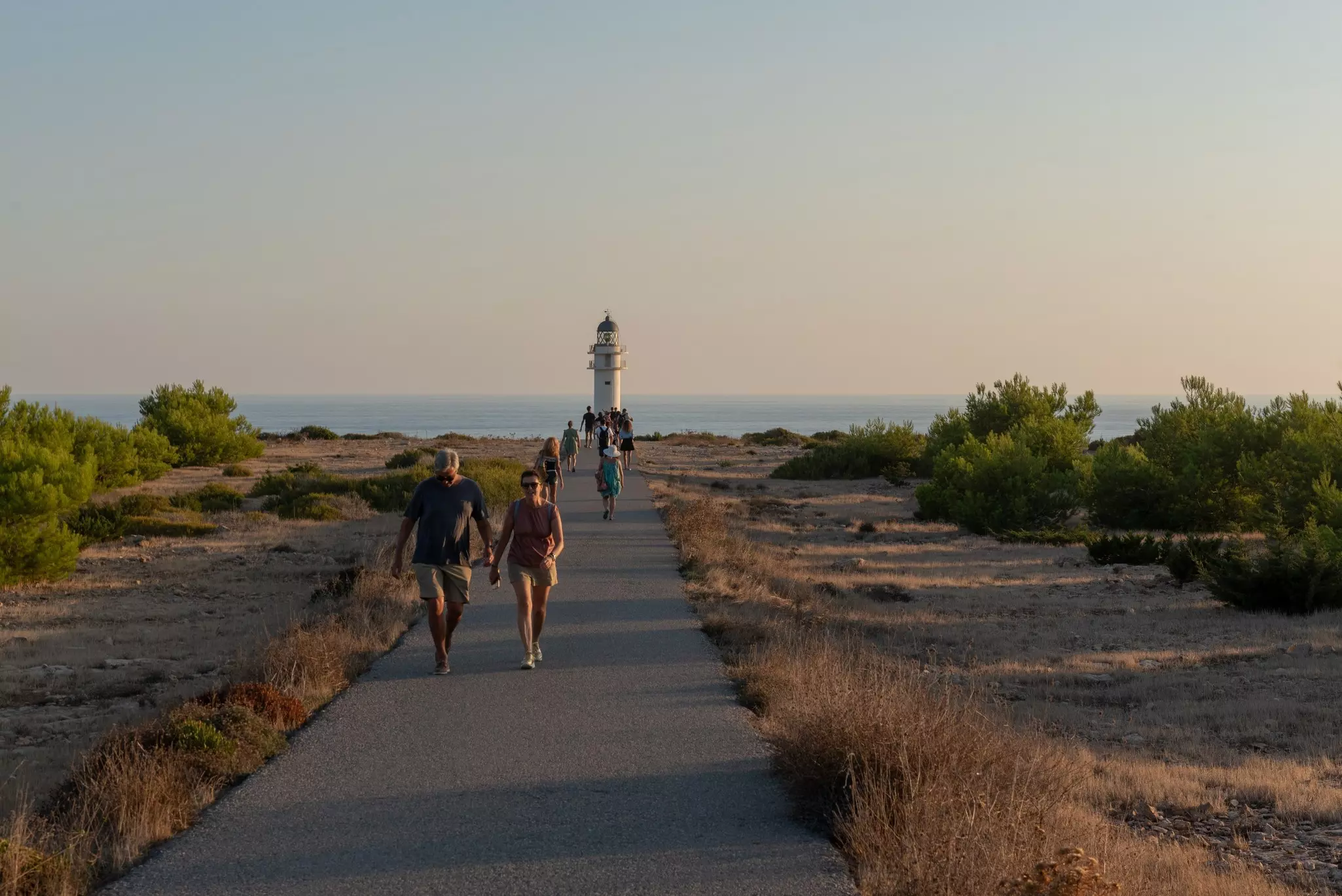 People walk along a path that leads to a lighthouse at dusk.