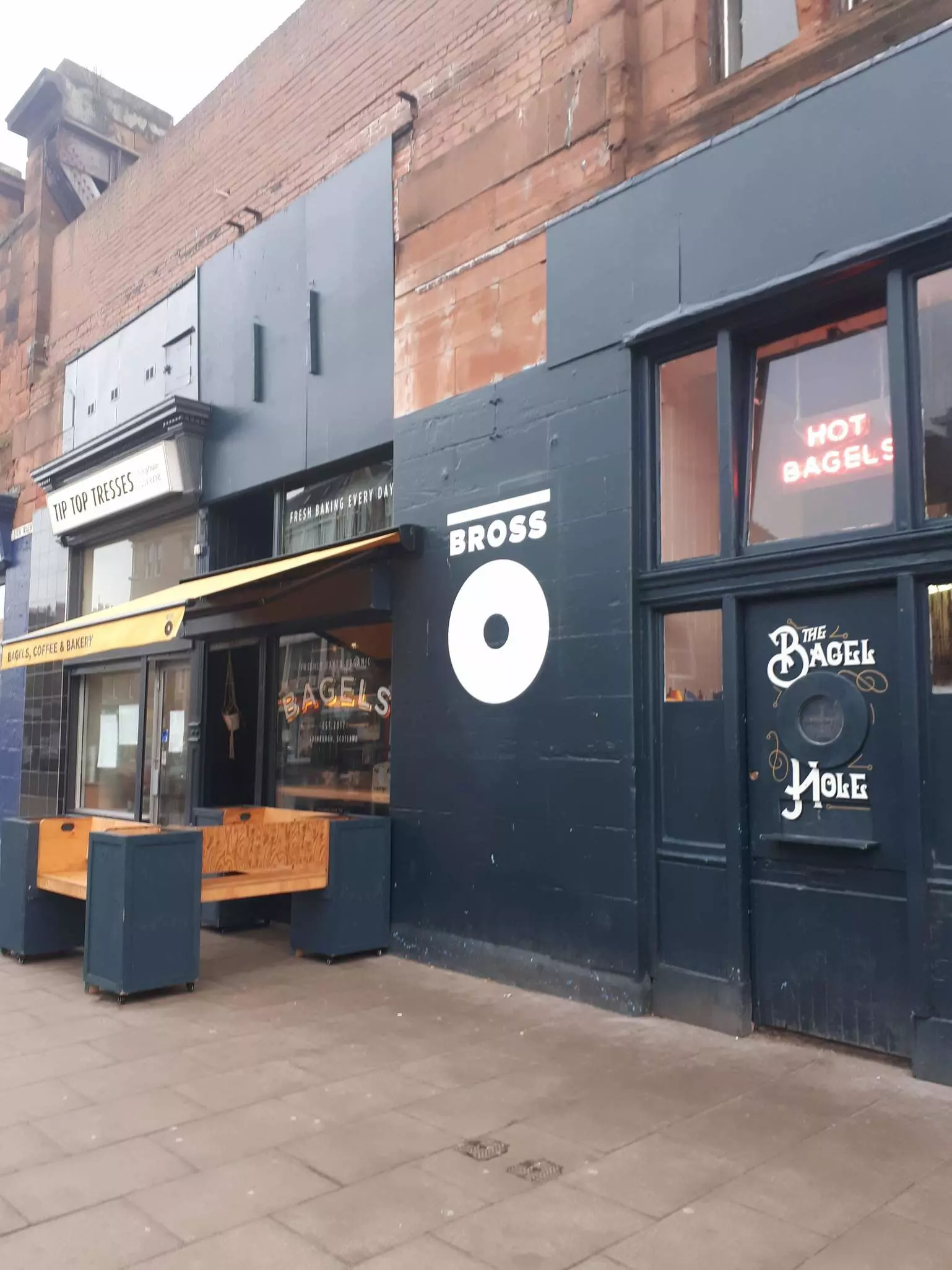 A close up of a hand holding a bagel, halved and wrapped in white grease-proof paper. The bagel is filled with cream cheese, capers, tomato and red onion.
