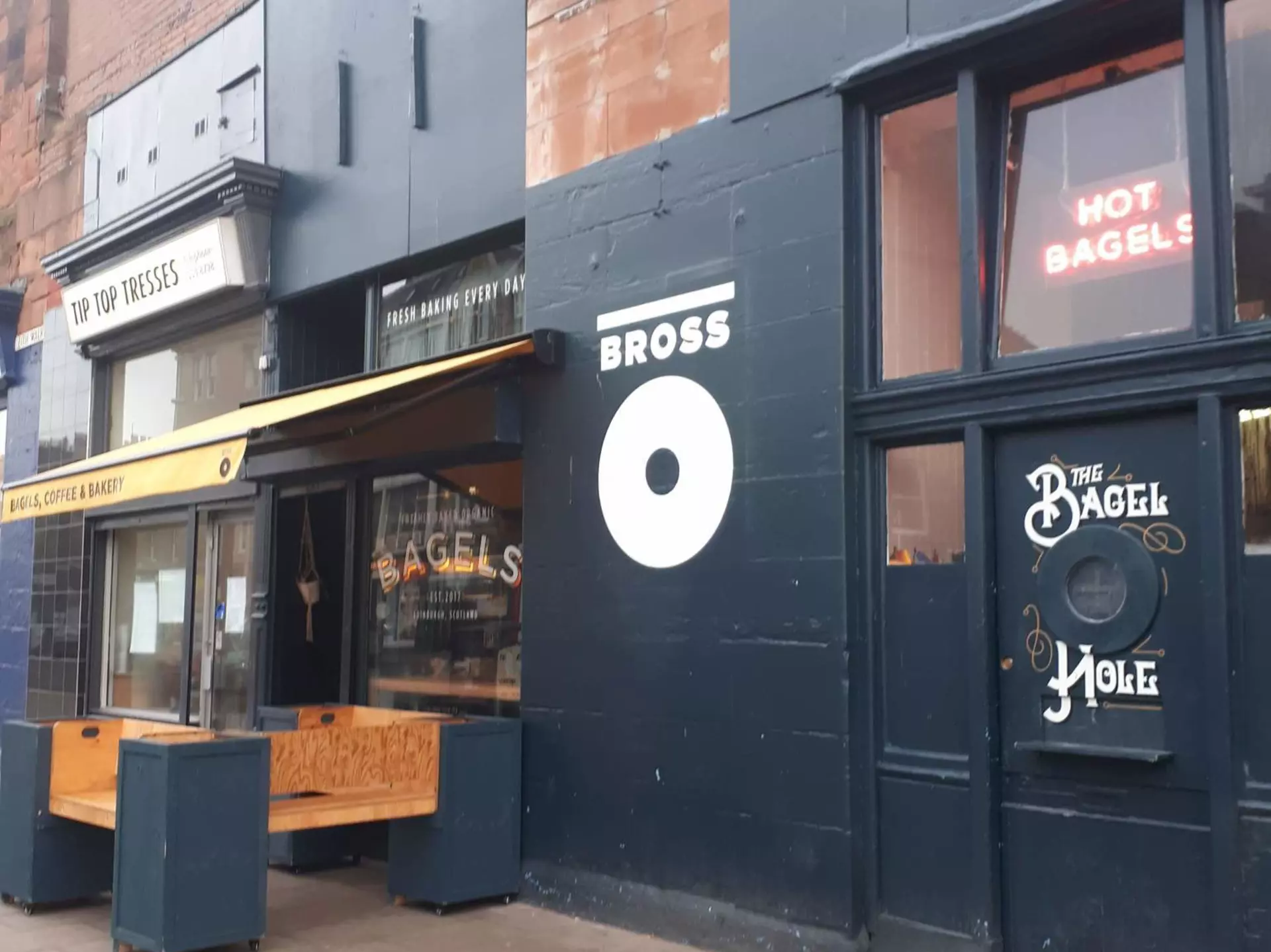 A close up of a hand holding a bagel, halved and wrapped in white grease-proof paper. The bagel is filled with cream cheese, capers, tomato and red onion.