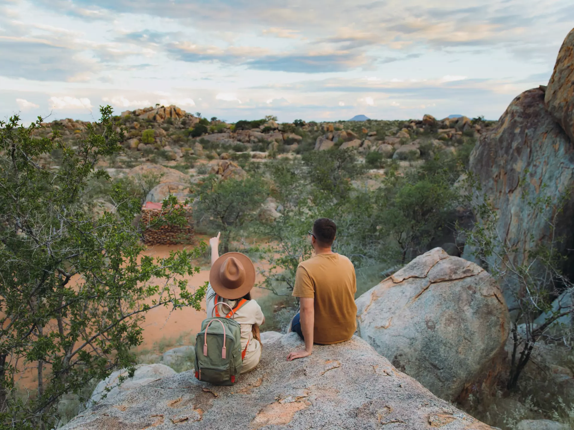 Young couple of woman in hat with backpack and man sitting on the top of the rock looking at the beautiful landscape and watching wild animals in Damaraland, Namibia, Southern Africa
