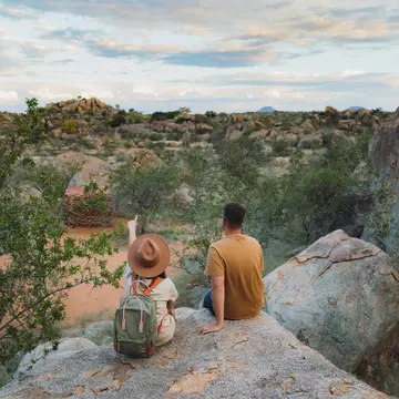 Young couple of woman in hat with backpack and man sitting on the top of the rock looking at the beautiful landscape and watching wild animals in Damaraland, Namibia, Southern Africa