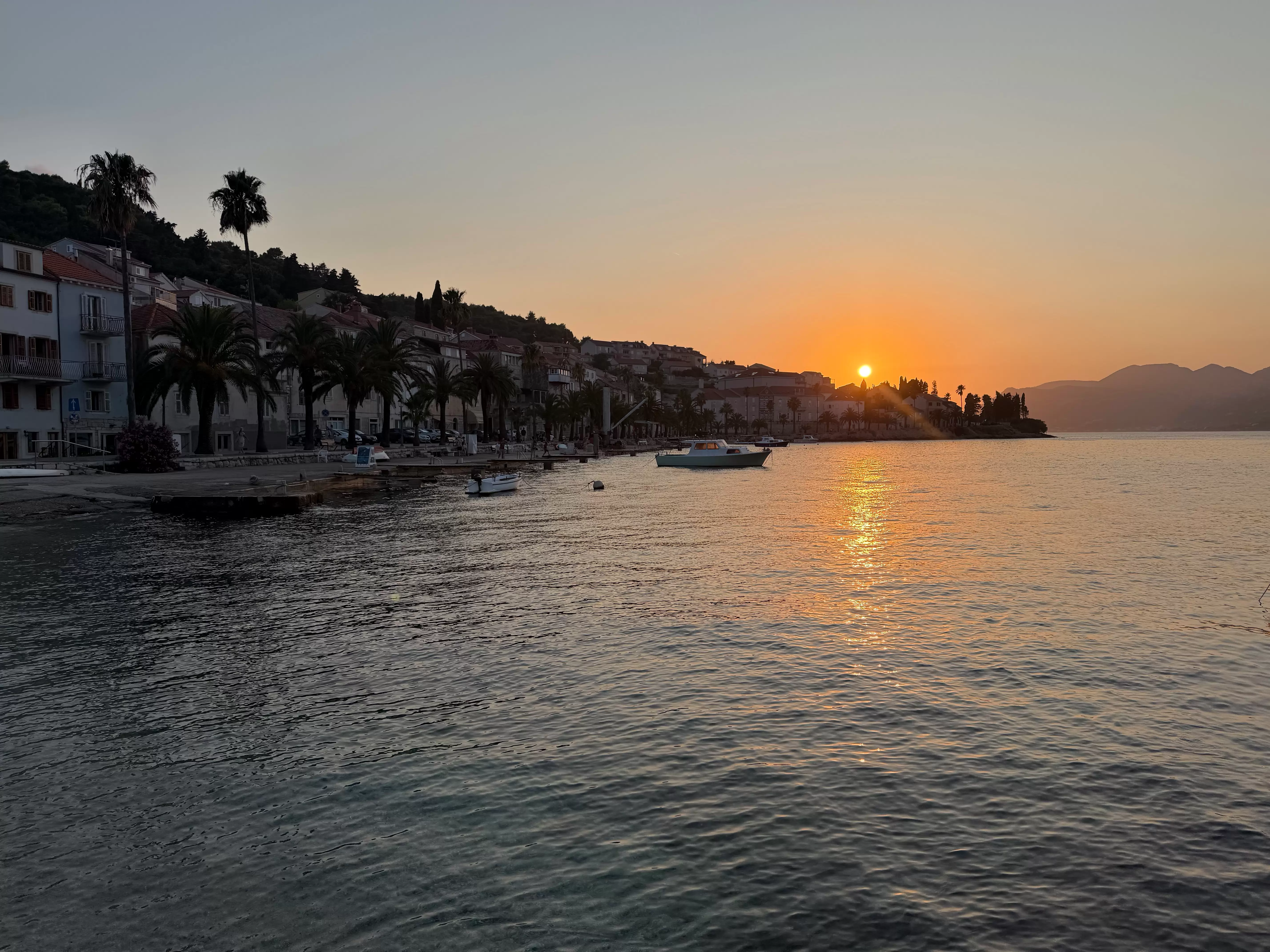The sun setting on the Adriatic Sea to the left a medium-sized ship is moored near the shore. There are palm dress and small businesses behind the boat.