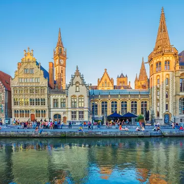Row of ornate gabled guildhouses and a spired Gothic tower on a Ghent quayside, people seated along the river's edge