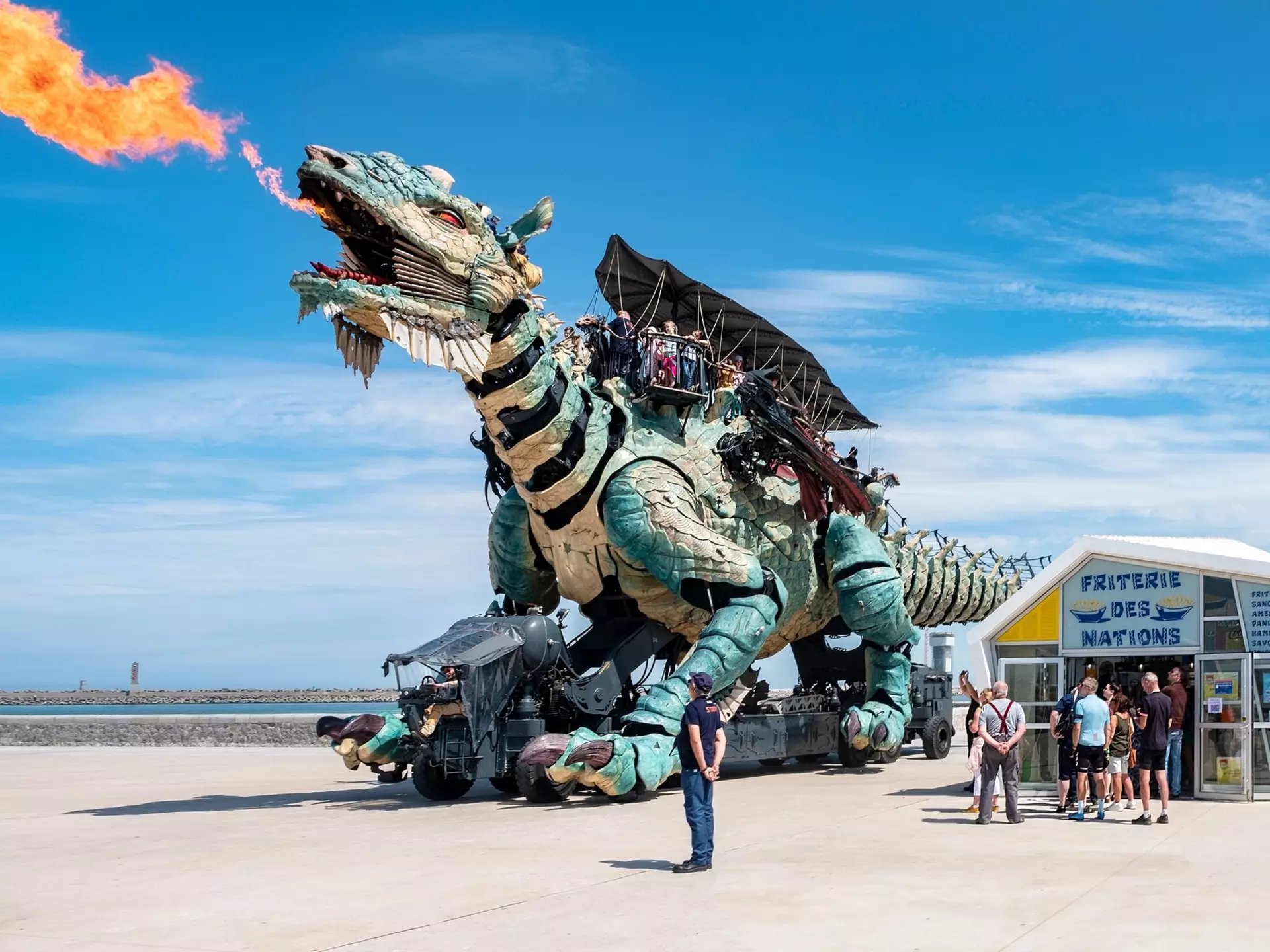 A giant mechanical dragon breathes fire near a beach with onlookers at ground level watching on