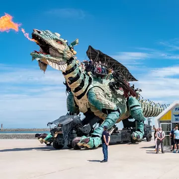 A giant mechanical dragon breathes fire near a beach with onlookers at ground level watching on