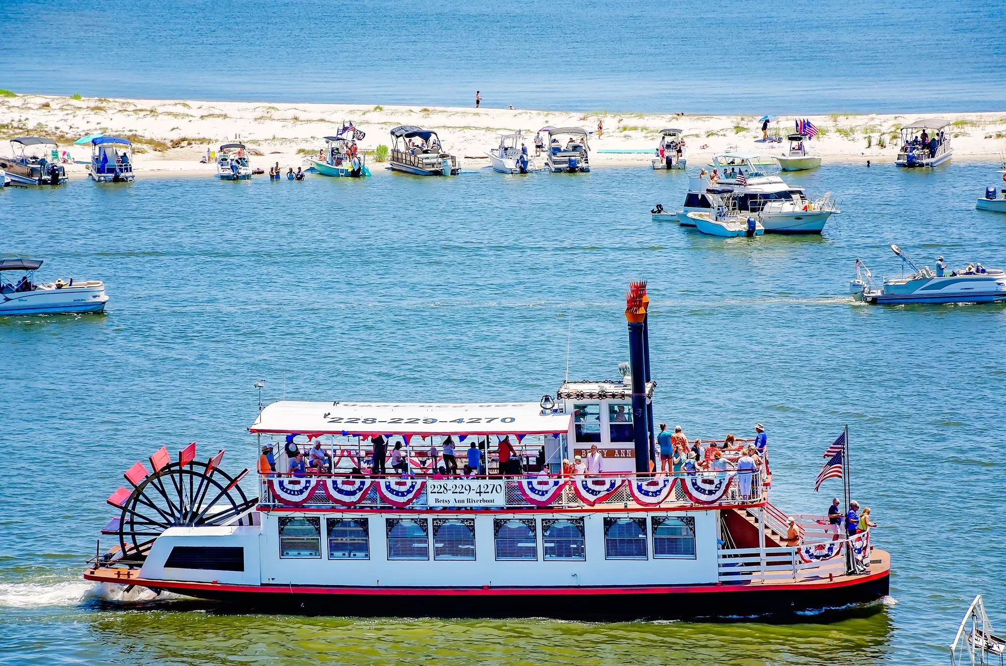 A riverboat with a large paddle passes other boats moored near a sandbar.