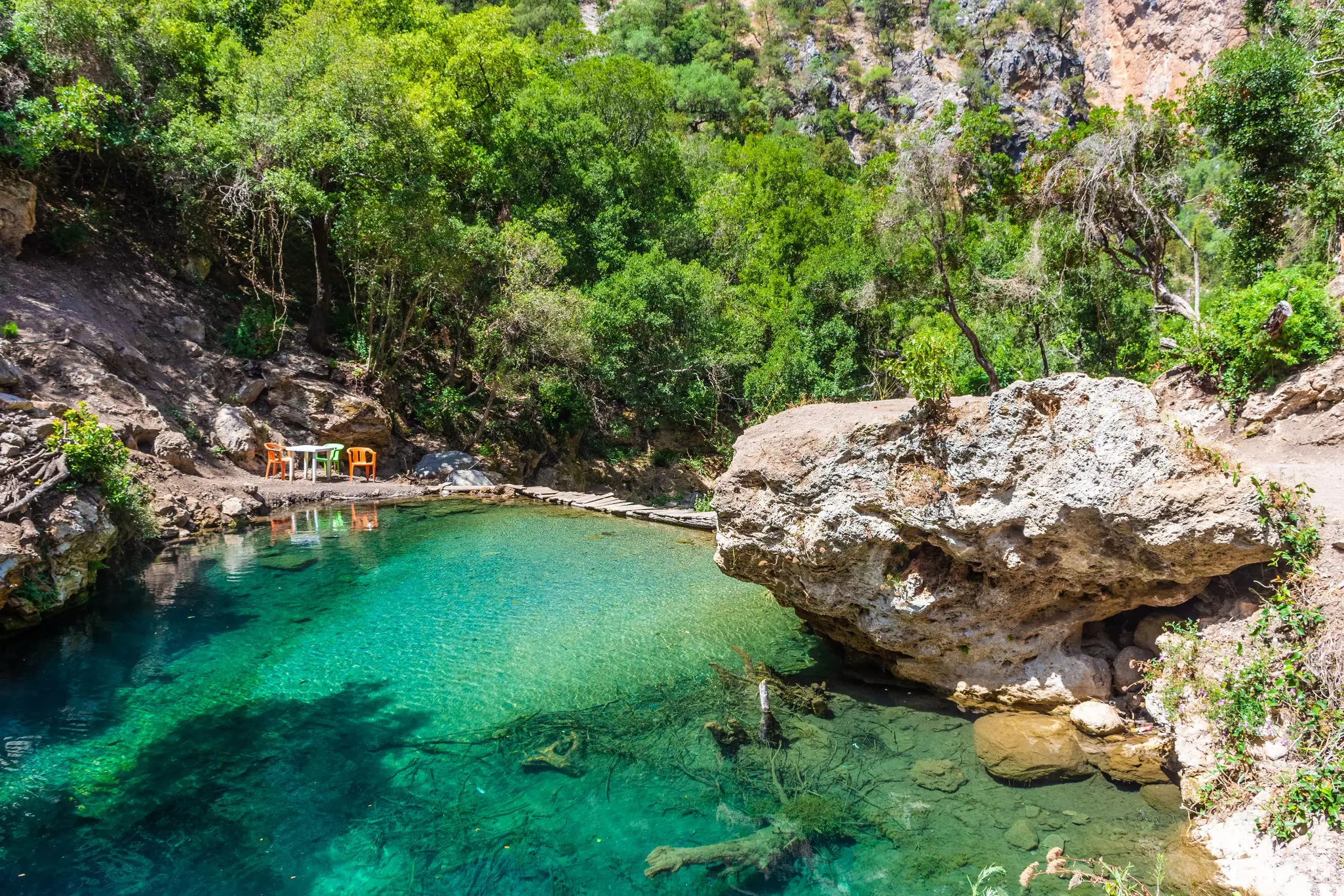 A very clear natural pool of green water, with a large rock on one shore and a plastic table with chairs set on a flat area on the other shore.