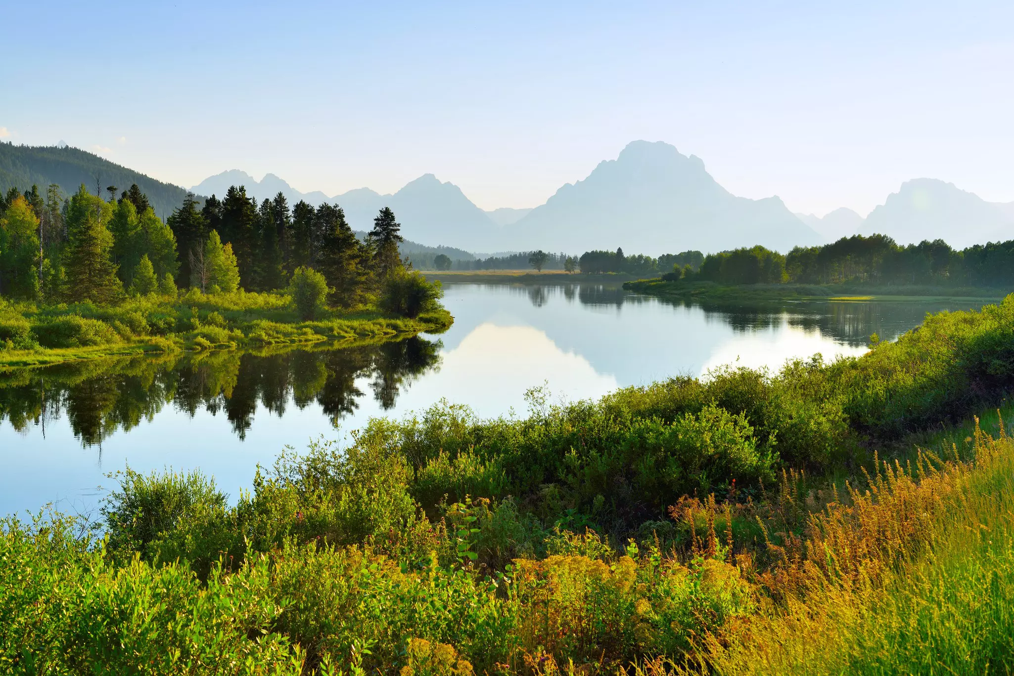 A still lake reflects distant mountains and is surrounded by lush green wetlands.