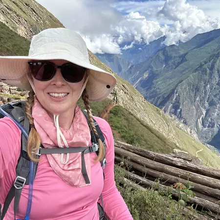 Woman in pink top and white hat and sunglasses smiling with mountainous scenery behind her