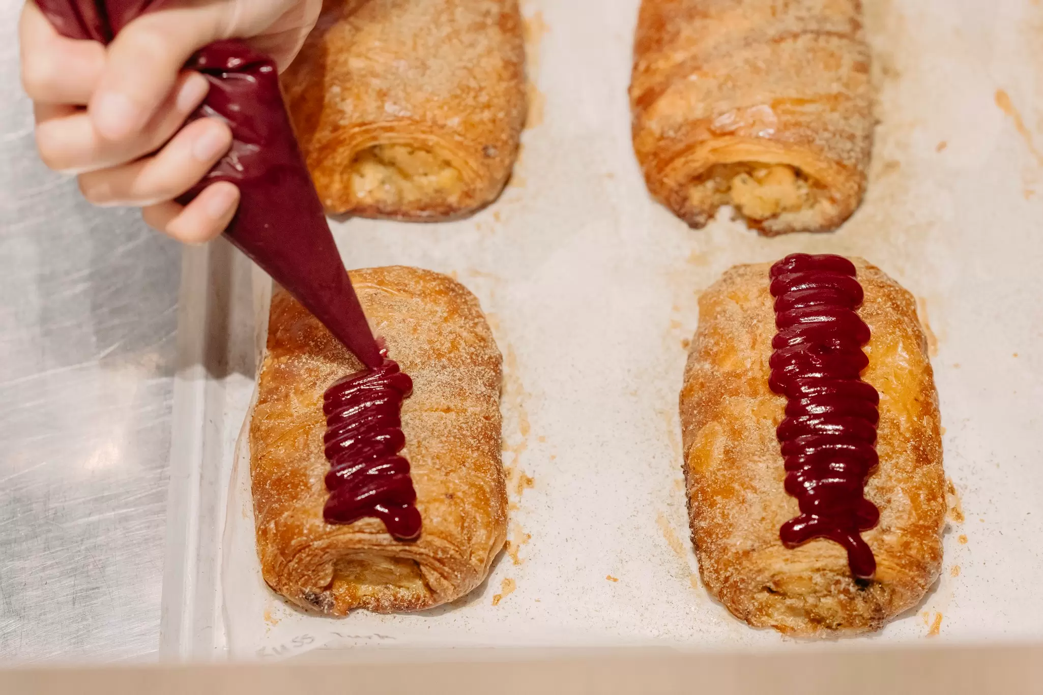 Four pastries arranged neatly. A person's hand with a frosting bag is visible, and the frosting is being squeezed onto one of the pastries.