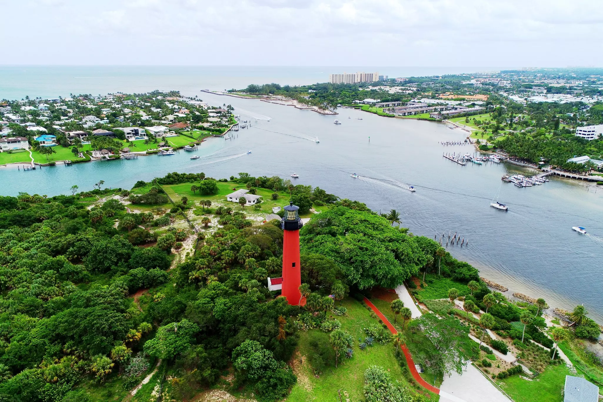 An aerial view of a red lighthouse on a small hill beside a canal that leads to the ocean. Boats are visible in the water, and many houses are seen among the green land.