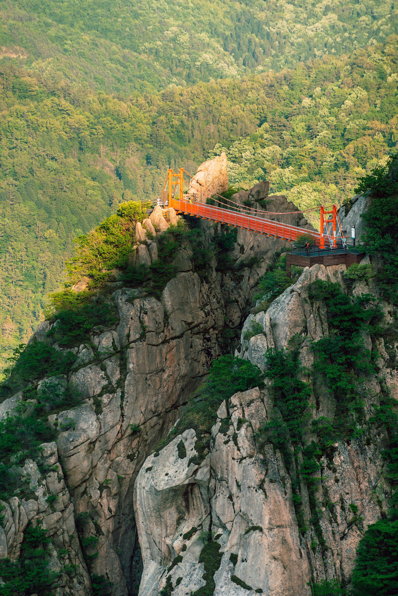 A red suspension footbridge between two rocky granite peaks above a forested valley in Wolchulsan National Park, South Korea.